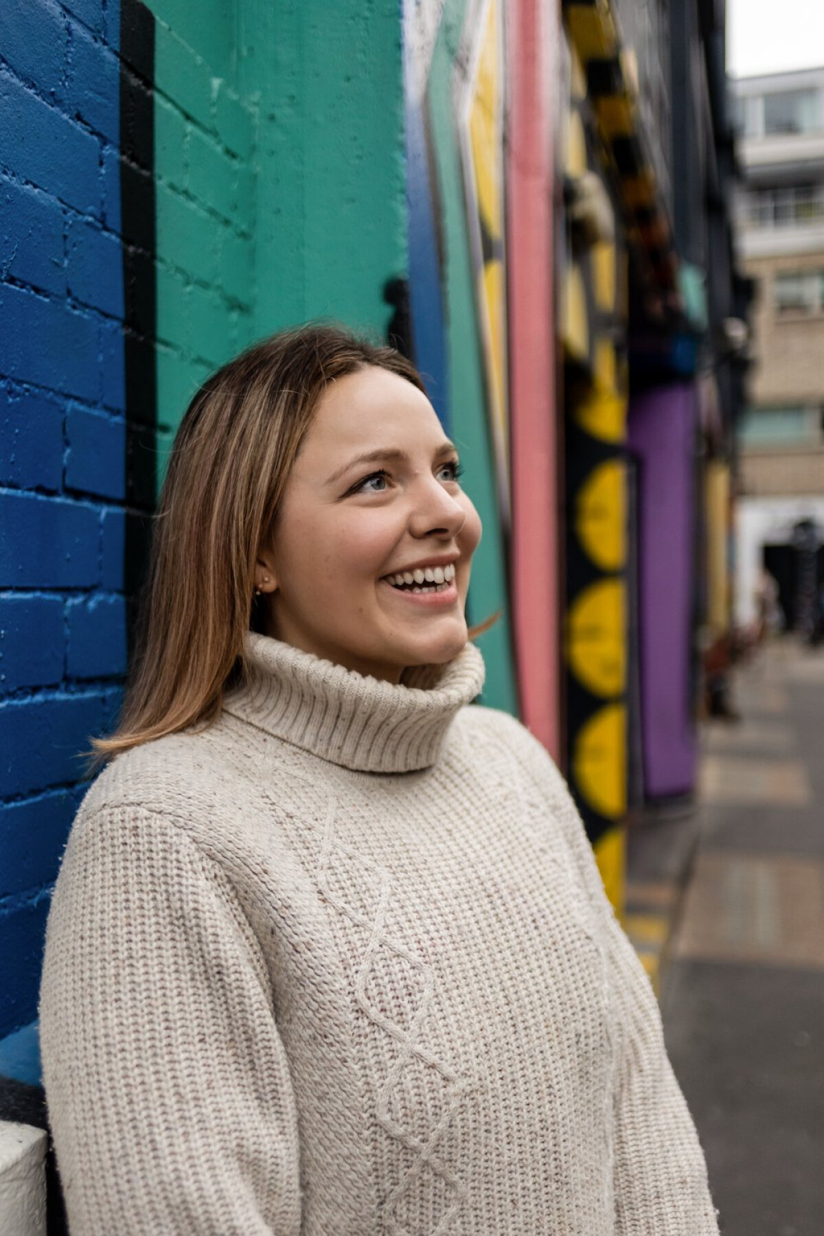 Ettie is a white person with light brown hair. She’s wearing a cosy beige jumper, and leaning against a colourful wall. She’s facing to the right, and smiling.